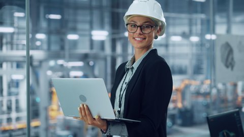Portrait of a Happy Young Beautiful Female Engineer Wearing White Hard Hat, Using Laptop Computer in Office at Car Assembly Plant. Industrial Specialist Working on Vehicle Design in Modern Facility.