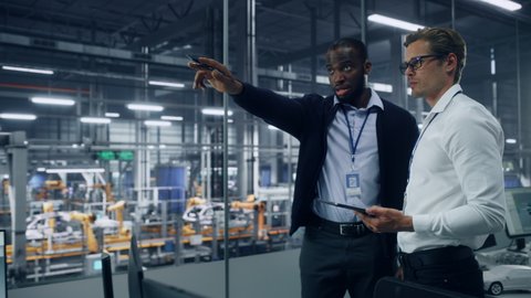 Two Diverse Automotive Industrial Engineers Talking About Vehicle Production while Standing in Office at a Car Assembly Plant. Industrial Specialists Discuss Work Projects on Tablet Computer.