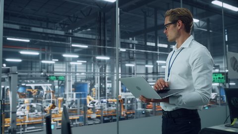 Handsome Engineer in Glasses and White Shirt Using Laptop Computer and Looking Out of the Office at a Car Assembly Plant. Industrial Specialist Working on Vehicle Design in Technological Facility.