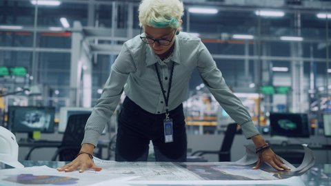 Young Female Engineer Looking at a Technical Blueprint at Work in an Office at Car Assembly Plant. Industrial Specialist Working on Vehicle Parts in Technological Development Facility.