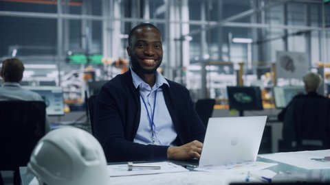 Portrait of a Happy Young African American Engineer Working on Laptop Computer in an Office at Car Assembly Plant. Industrial Specialist Working on Vehicle Parts in Technological Development Facility.