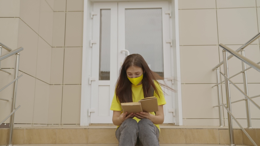 A schoolgirl girl in a mask reads a book while standing at the school, protection against coronavirus, covid-19 pandemic, air filtration through a mask filter, modern education for adolescents Royalty-Free Stock Footage #1077851186