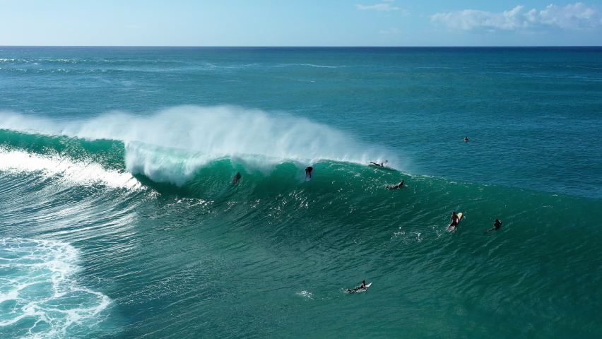 Aerial Tracking A Surfer Riding A Giant Breaking Wave With Bright Sunlight, White Foaming Surf, And Other Surfers Paddling Nearby - Oahu, Hawaii