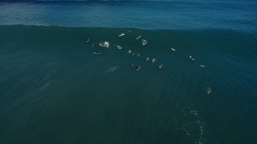 Aerial Tracking An Intent Surfer Riding A Giant Crashing Wave With Deep Blue Ocean Water, White Foaming Surf, And Other Surfers Paddling Nearby - Oahu, Hawaii