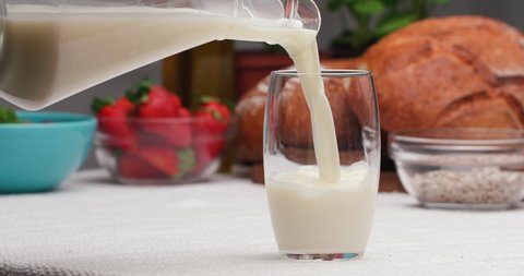 Fresh Milk is Pouring from a Jug into Glass on the table with Breakfast Food. Filling Drinking Glass with Milk at Breakfast Time. Concept of Healthy Dairy Product. Strawberries and Bread on Background