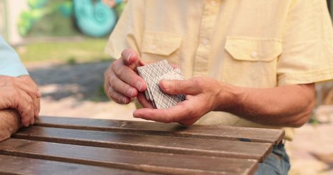 Cropped portrait shot of the caucasian senior man sitting at the table outdoors in the yard, playing cards game and talking. Man shuffles a deck of cards