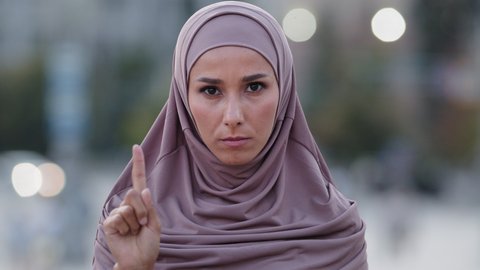 Female portrait outdoors in city serious islamic woman muslim dissatisfied girl wears hijab looking at camera shakes index finger no saying not never denies forbids disapproves waving head negative