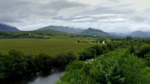 Bealanabrack River, Maum, Connemara, County Galway, Ireland, July 2021. Drone follows the course of the river west over grassy marshland while slowly ascending and revealing Maumturk Mountains.