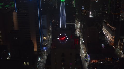 Backwards reveal and tilt up of neon illuminated spike and clock on top of Mercantile National Bank Building. Aerial drone night view. Dallas, Texas, US in 2021