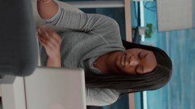 Vertical video: Student blogger with dark skin putting headphones working from home at social media articole browsing communication lecture on computer. African teenager sitting on bean bag solving e