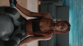 Verical video: Fit woman with dark skin doing exercises with fitness dumbbells sitting on swiss ball working at morning workout aerobic in living room practicing sport. Adult with sportswear training