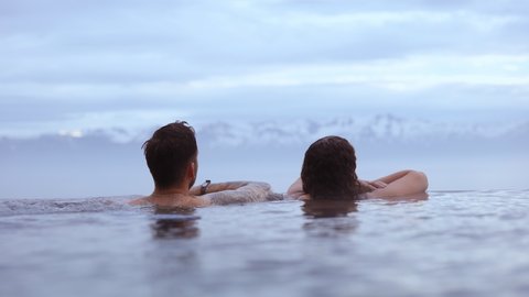 REYKJAVIK, ICELAND - JUNE 2021: Steady zoom-in shot of a couple dipped on a hot infinity pool, relaxing with view of majestic mountains with snow on its peak beneath the brilliant blue heavens