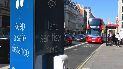 London, UK - July 1, 2021: a COVID sign asking people to social distance and hand sanitise next to a road in London, with a bus stop in the background, as a Red London Double Decker Bus drives past