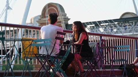 London, UK - July 1, 2021: a couple enjoying a drink next to the River Thames as the sun sets on the South Bank. Behind there is Hungerford Bridge and the Golden Jubilee Bridges. A train passes over
