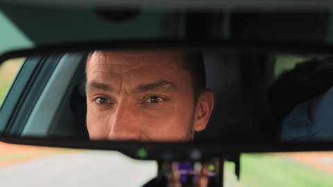 Close-up eyes of a young man are reflected in the rear-view mirror of a car. The guy looks at the road, then at the camera. The concept of travel and recreation by car on vacation or weekend. UHD 4K.