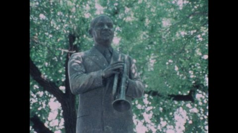 1970s: Statue of William Christopher Handy in park. Plaque on statue. People pose for photo at gates to Graceland. Marquee outside Stax Records. Man plays saxophone on stage with Isaac Hayes.