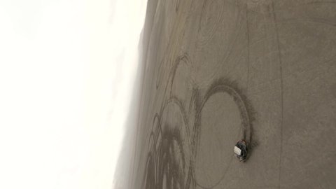 Large buggy automobiles drift on dried salt lake territory with brown sand leaving circle traces against hill silhouettes. Aerial top view on freestyle fpv drone
