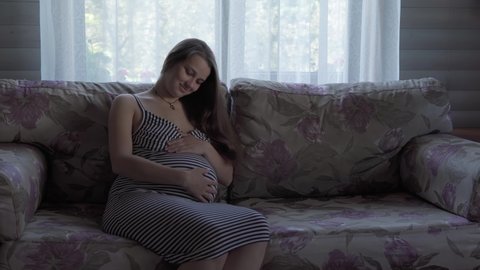 cute happy tired pregnant woman touching her tummy resting sitting in bed at home. caring mother take rest stroke belly on light provence style room. pregnancy, motherhood, people, expectation concept