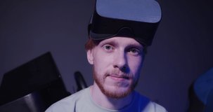 A red-haired guy of European appearance in virtual reality glasses sits on a virtual reality simulator and seriously looks straight into the camera