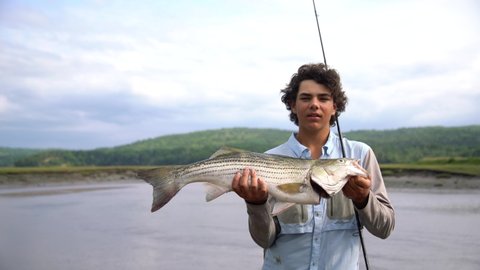 A young sport fisherman holding a nice striped bass by a tidal river in Nova Scotia, Canada. Real scenario.