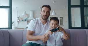 Father hugging his son while actively playing on computer games on joystick, looking at the screen and expressing happiness and excitation when winning. 4K video footage