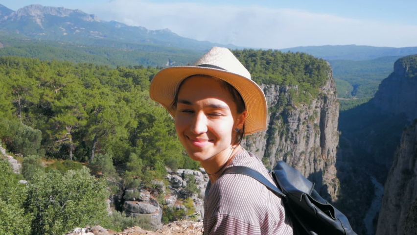 A young woman traveler in a white hat and with a backpack on her shoulders stands on the top of a cliff and looks at the mountain landscape. A young woman hiker has reached the mountain peak and