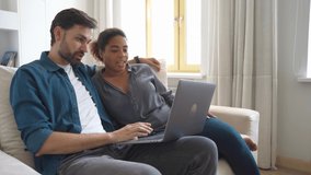 Happy interracial couple looking at laptop screen sitting on sofa at home. Multiracial family shopping online together. Black woman caucasian man in love using computer watching video surfing internet