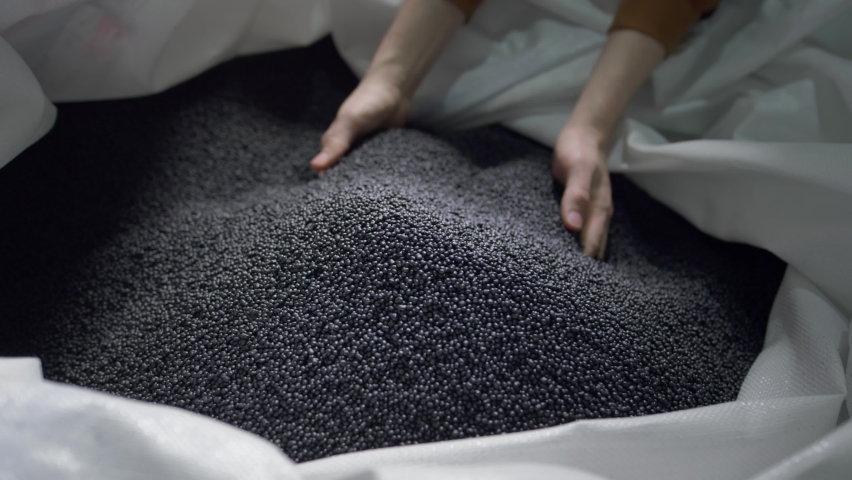Hands lift black polymer granules from a cloth bag in a garbage recycling plant. Plastic pellets are crumbling or poured from the palms. Raw materials for recycled plastic are used in manufacturing. | Shutterstock HD Video #1077949790