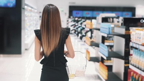 Back view of young Caucasian woman walks in cosmetics store. Slim brunette looks for some cosmetic and walks past stalls with shopping basket. Concept of shopping. High quality 4k footage.