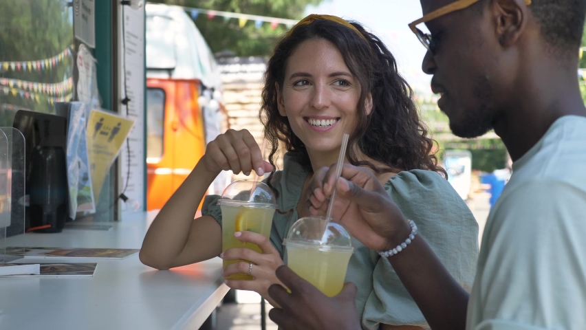 Afro-American man and young white woman drink lemonade with swizzle sticks smiling near kiosk in attraction park on sunny day closeup