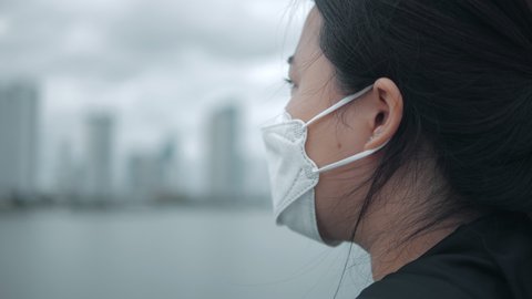 Close up of tired Asian woman wearing face mask standing outdoors with a serious expression thinking against view of the city skyline with looming dark clouds