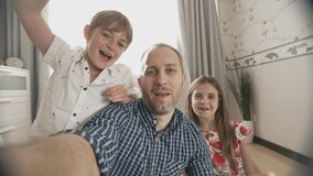 Selfie portrait of family of three smiling and laughing to camera. Wide angle view shot of father, son and daughter video chats on smart phone on living room background.