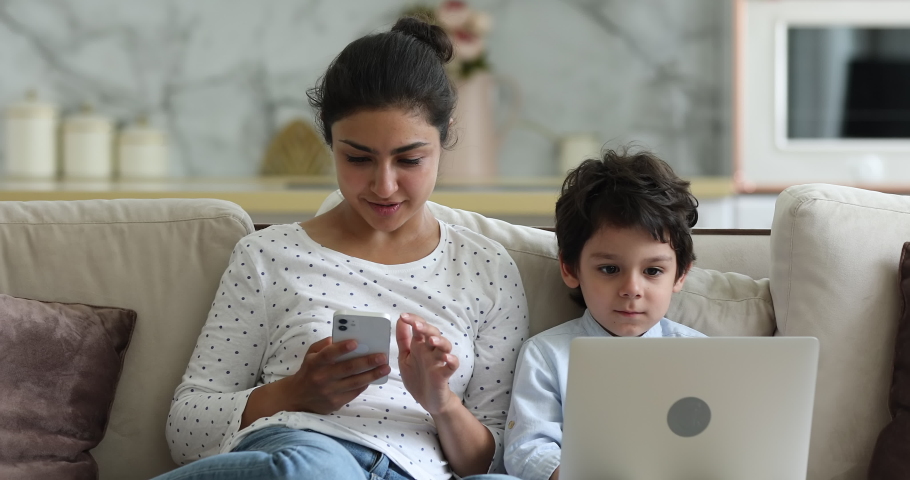 Indian woman her little son sit on sofa use gadgets. Mom hold phone 4s boy put laptop on laps enjoy online fun play video games. Devices overuse, young generation internet technology addiction concept Royalty-Free Stock Footage #1077958214