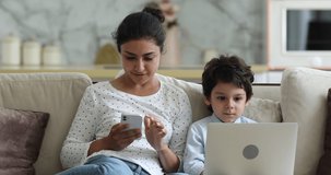 Indian woman her little son sit on sofa use gadgets. Mom hold phone 4s boy put laptop on laps enjoy online fun play video games. Devices overuse, young generation internet technology addiction concept