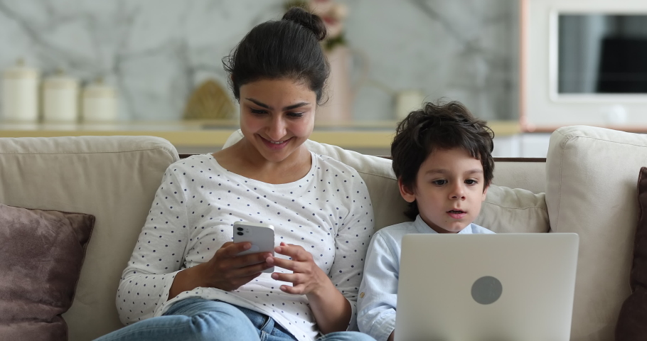 Indian woman her little son sit on sofa use gadgets. Mom hold phone 4s boy put laptop on laps enjoy online fun play video games. Devices overuse, young generation internet technology addiction concept | Shutterstock HD Video #1077958214