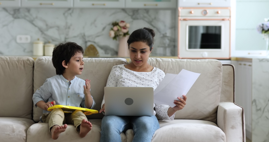 Little boy makes noise disturbs, distracting Indian freelancer mom try to work, do remote paperwork use laptop sit on sofa with kid feels irritated due restless son need attention. Lockdown concept | Shutterstock HD Video #1077958217