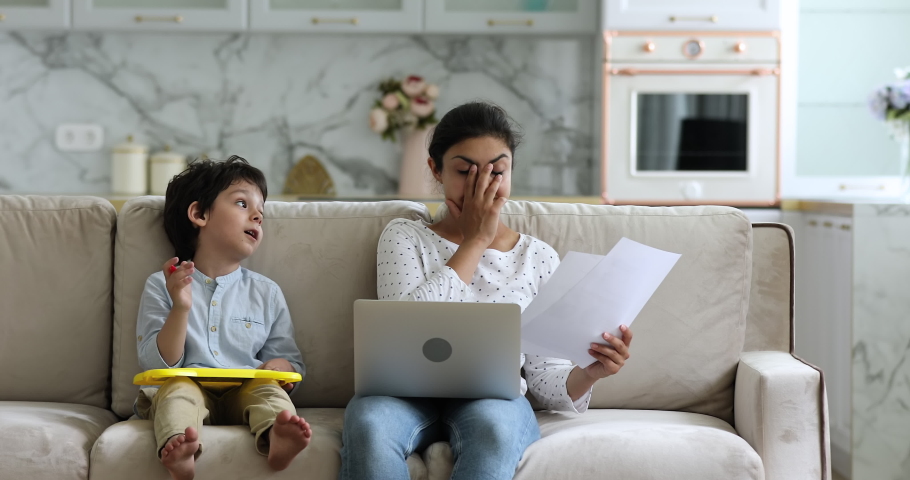 Little boy makes noise disturbs, distracting Indian freelancer mom try to work, do remote paperwork use laptop sit on sofa with kid feels irritated due restless son need attention. Lockdown concept | Shutterstock HD Video #1077958217