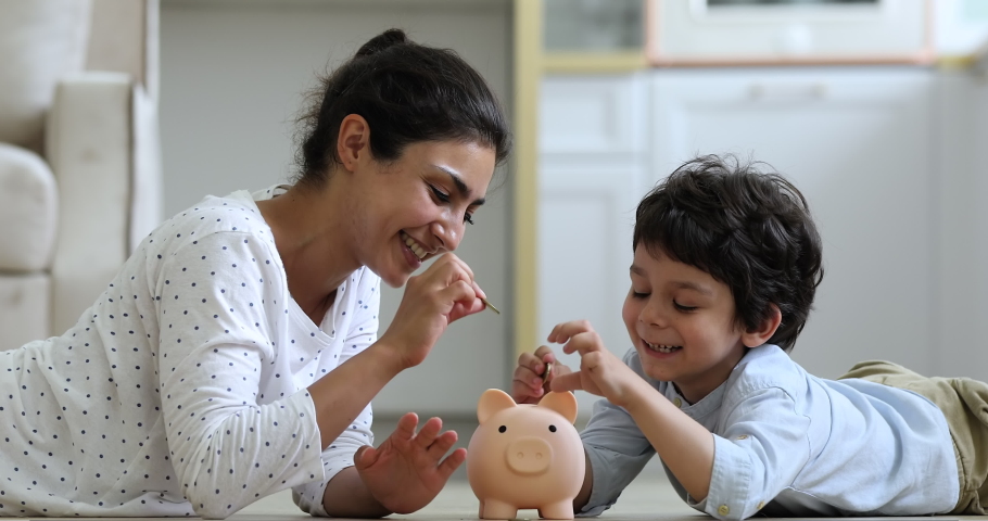 Young Indian mother her little 4s son lying on floor at home count and drop coins into piggy bank. Caring parent teaches child save money, think about future, manage personal finances, savings concept