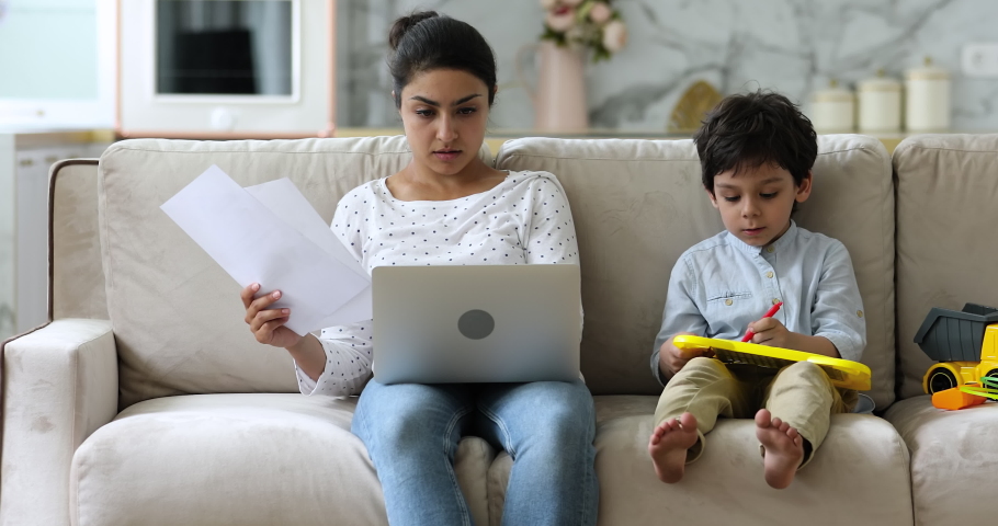 Busy concentrated Indian entrepreneur woman do remote paperwork accounting job use laptop working sit on sofa with little son drawing on magnetic erasable board. Quarantine at home with kids concept Royalty-Free Stock Footage #1077958385