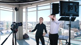 Young bloggers recording video about their business. Creative young couple shooting video in front of professional camera in office with panoramic windows.
