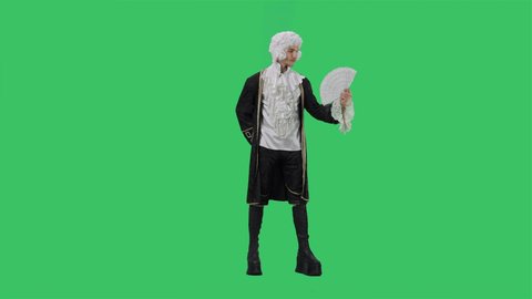 Portrait of courtier gentleman in black historical vintage suit and white wig, dancing merrily with fan in his hands. Young man posing in studio with green screen background. Full lenght. Slow motion.