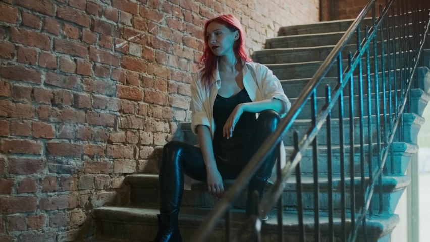 A beautiful red-haired girl wearing leather trousers, belts, black and white top and ring in her nose looking straight sitting on stairs in red brick building or block of flats Royalty-Free Stock Footage #1077961139