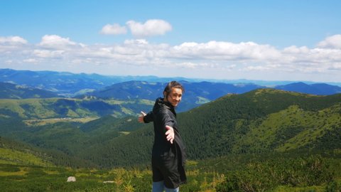 A young attractive woman traveler stands on the top of the mountain and looks into the distance at the majestic forest. A young woman traveler stands on the top of the cliff and looks at the mountain