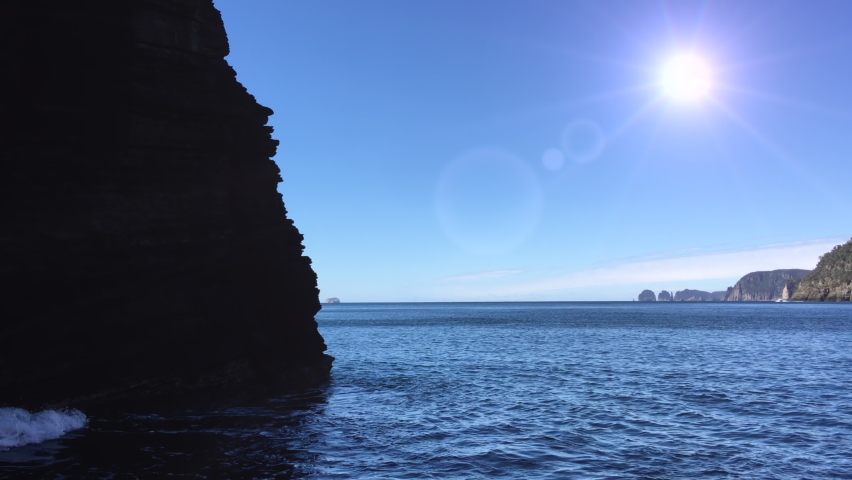 Sun and sea. Tracking past silhouette of rocky outcrop. Ocean swells lap at sandstone cliffs, silhouetted against bright sun beating down. Tasman Peninsula, Tasmania. Australia.