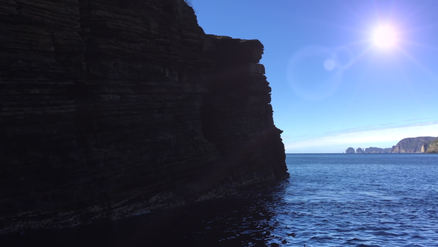 Sun and sea. Tracking past silhouette of rocky outcrop. Ocean swells lap at sandstone cliffs, silhouetted against bright sun beating down. Tasman Peninsula, Tasmania. Australia.