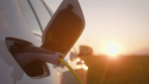Close up shot of an opened electric car charging socket cap and charger plugged into, at a public electric charging station near the highway at sunset