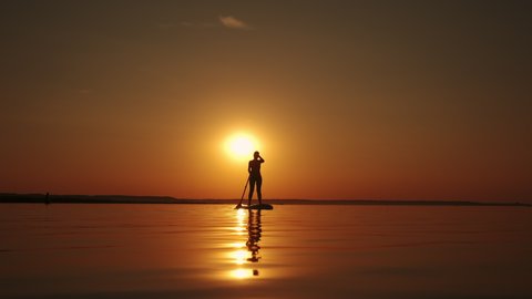 Siluet of Woman standing firmly on inflatable SUP board and paddling through shining water surface. Wide shot