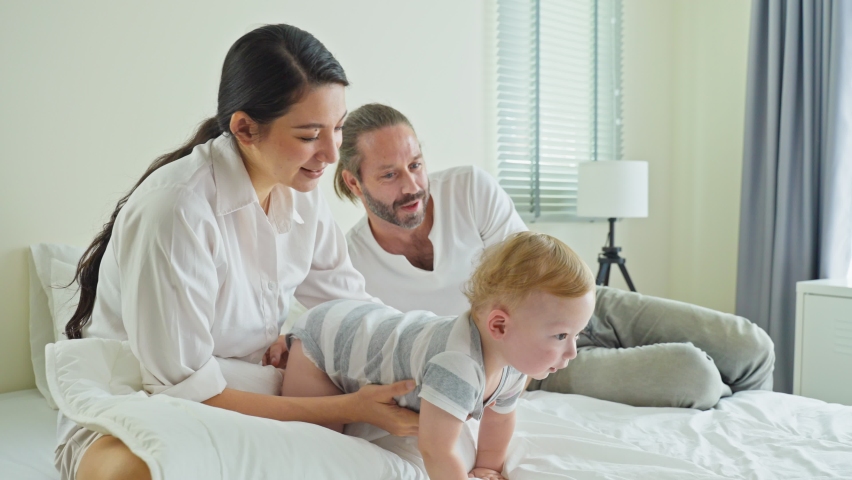 Caucasian loving parents play with baby boy child on bed in bedroom. Happy family, attractive beautiful caring young mother  and father spend time with toddler son crawling on bed in morning in house.