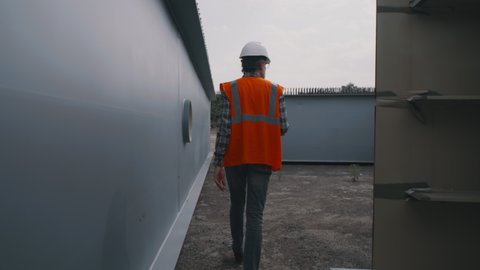 Tracking shot of middle aged man in waistcoat and hardhat walking near metal parts while working on construction site