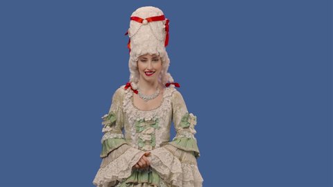 Portrait of courtier lady in white vintage dress and wig smiling coquettishly, making eyes, winking. Young woman posing in studio with blue screen background. Close up. Slow motion ready 59.94fps.